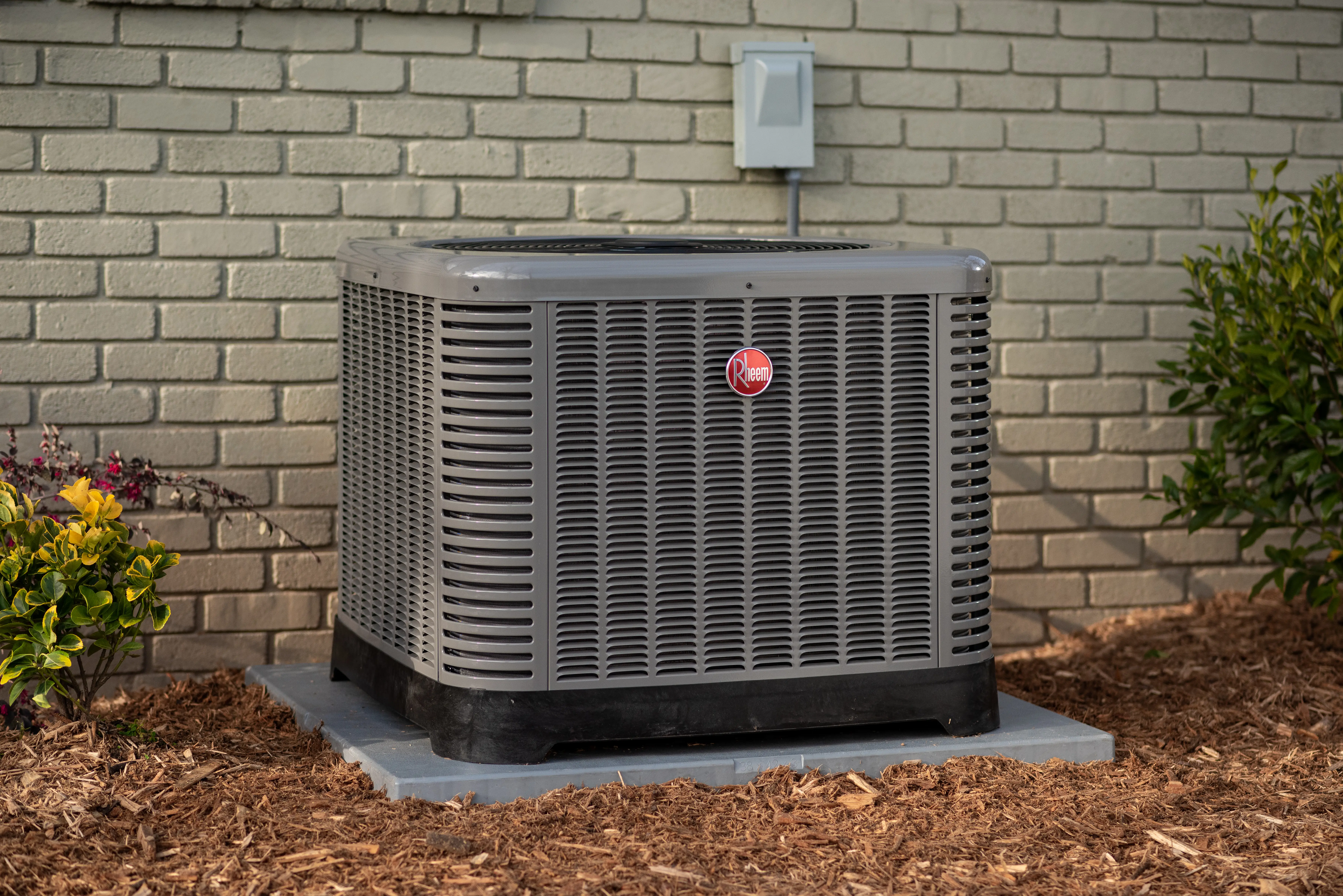 A Rheem air conditioning unit is shown outdoors, situated on a concrete pad next to a brick wall. The unit is gray with a black base and a red Rheem logo. Landscaping includes mulch, small bushes, and a gray electrical box mounted on the brick wall.