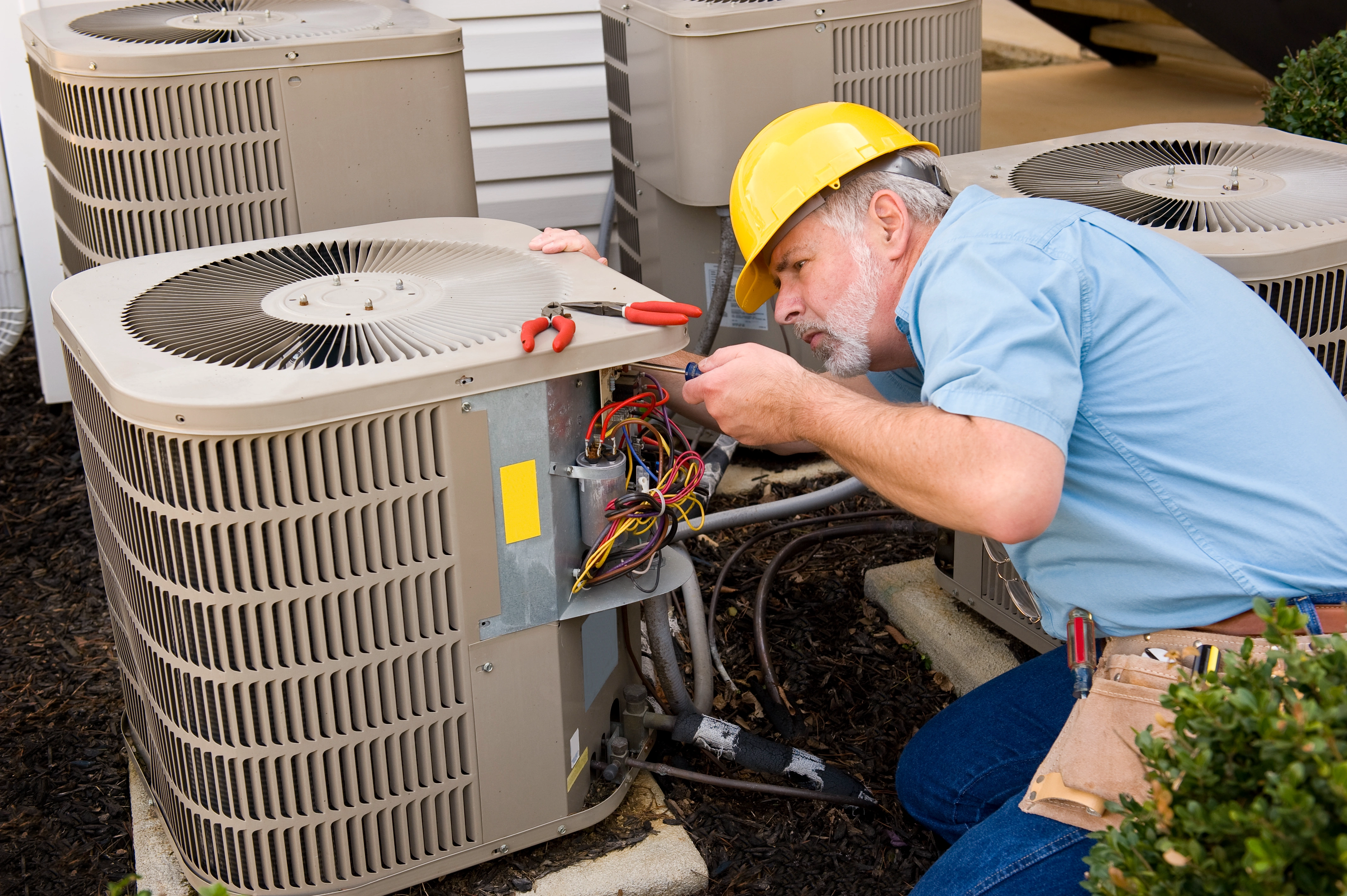 A technician in a yellow hard hat and blue shirt repairs an outdoor air conditioning unit.