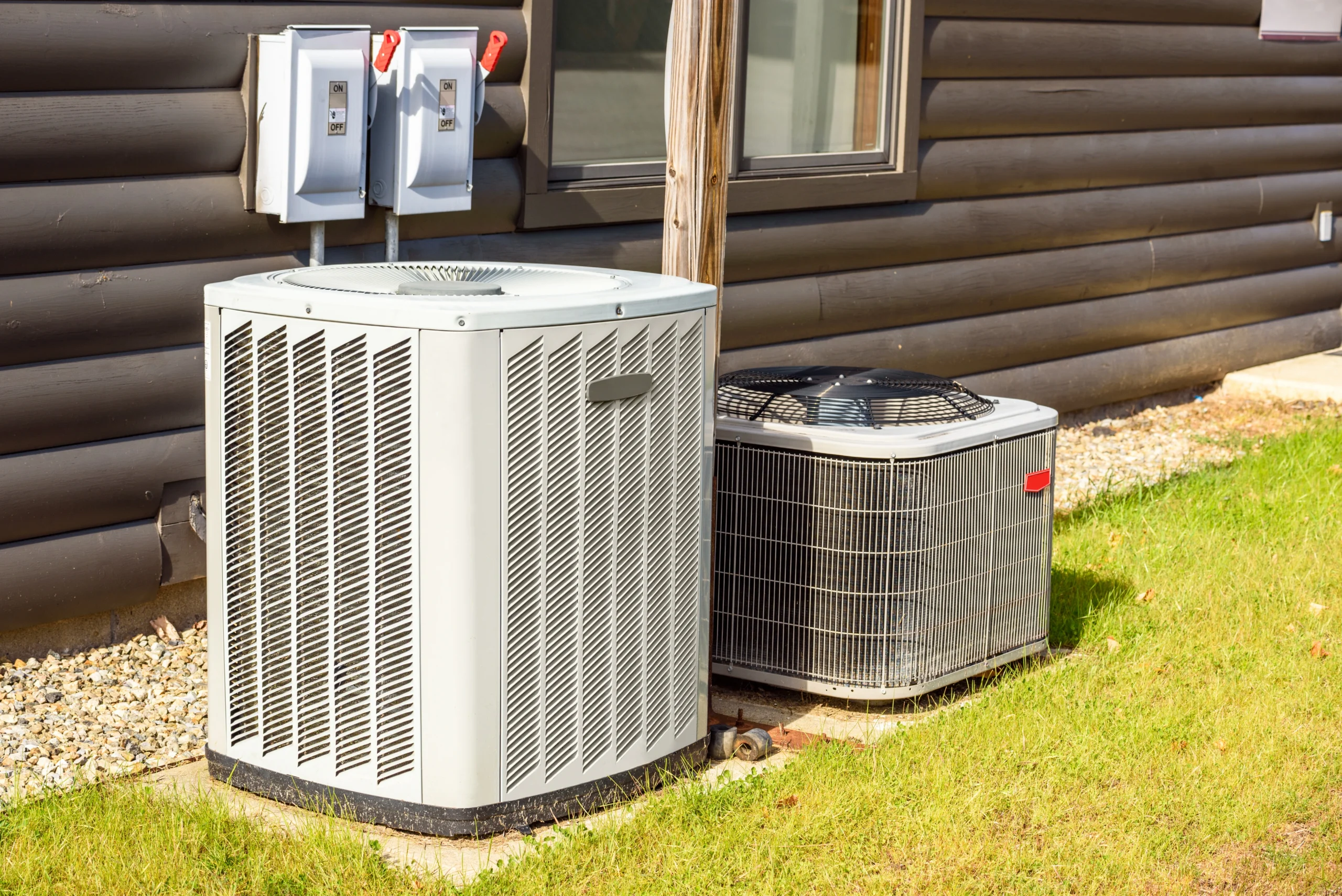 Two outdoor air conditioning units are installed on concrete pads next to a house with dark wood siding.