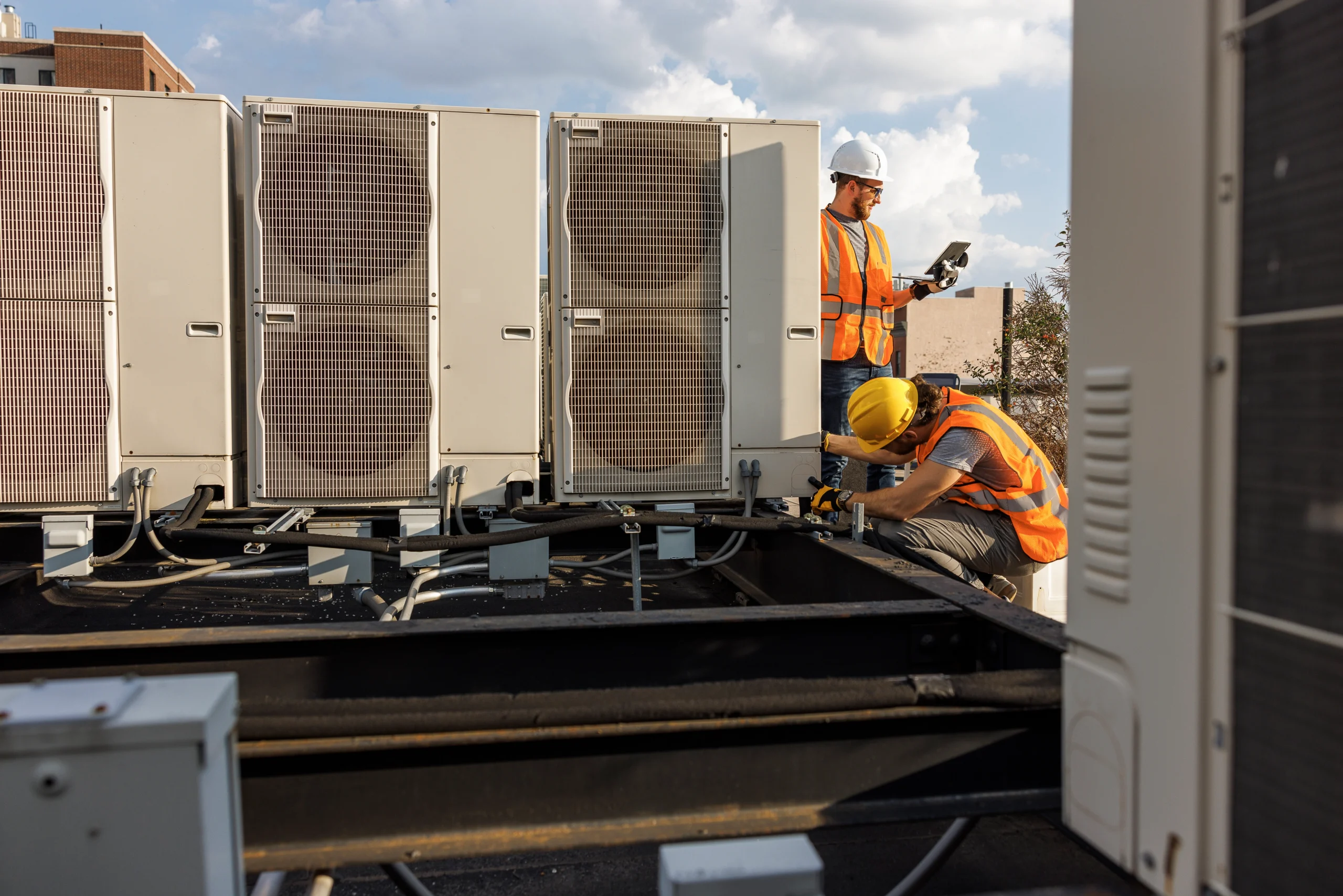 Two construction workers in safety gear inspect rooftop HVAC units on a sunny day.