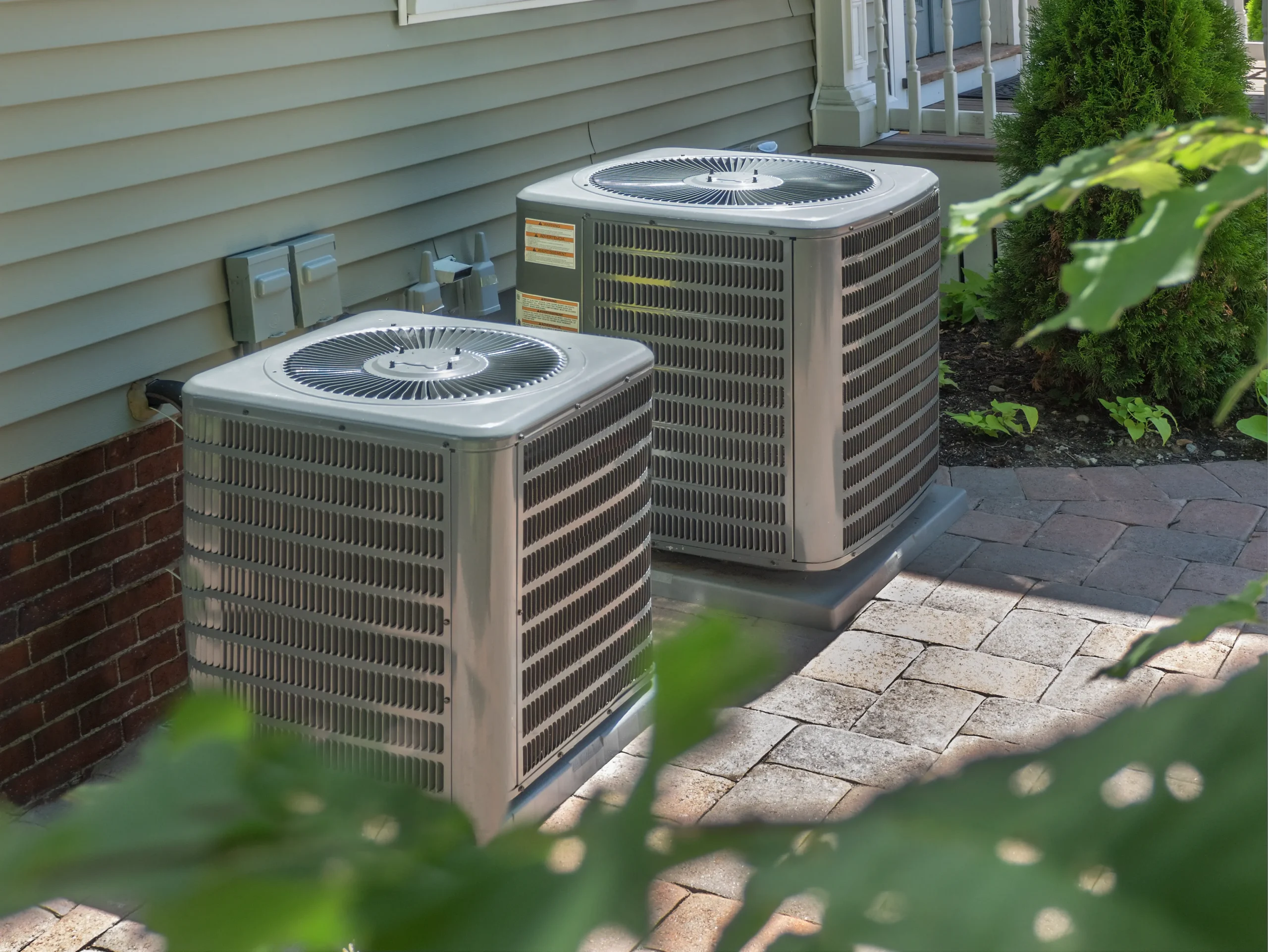 Two outdoor air conditioning units on a brick patio beside a house. 