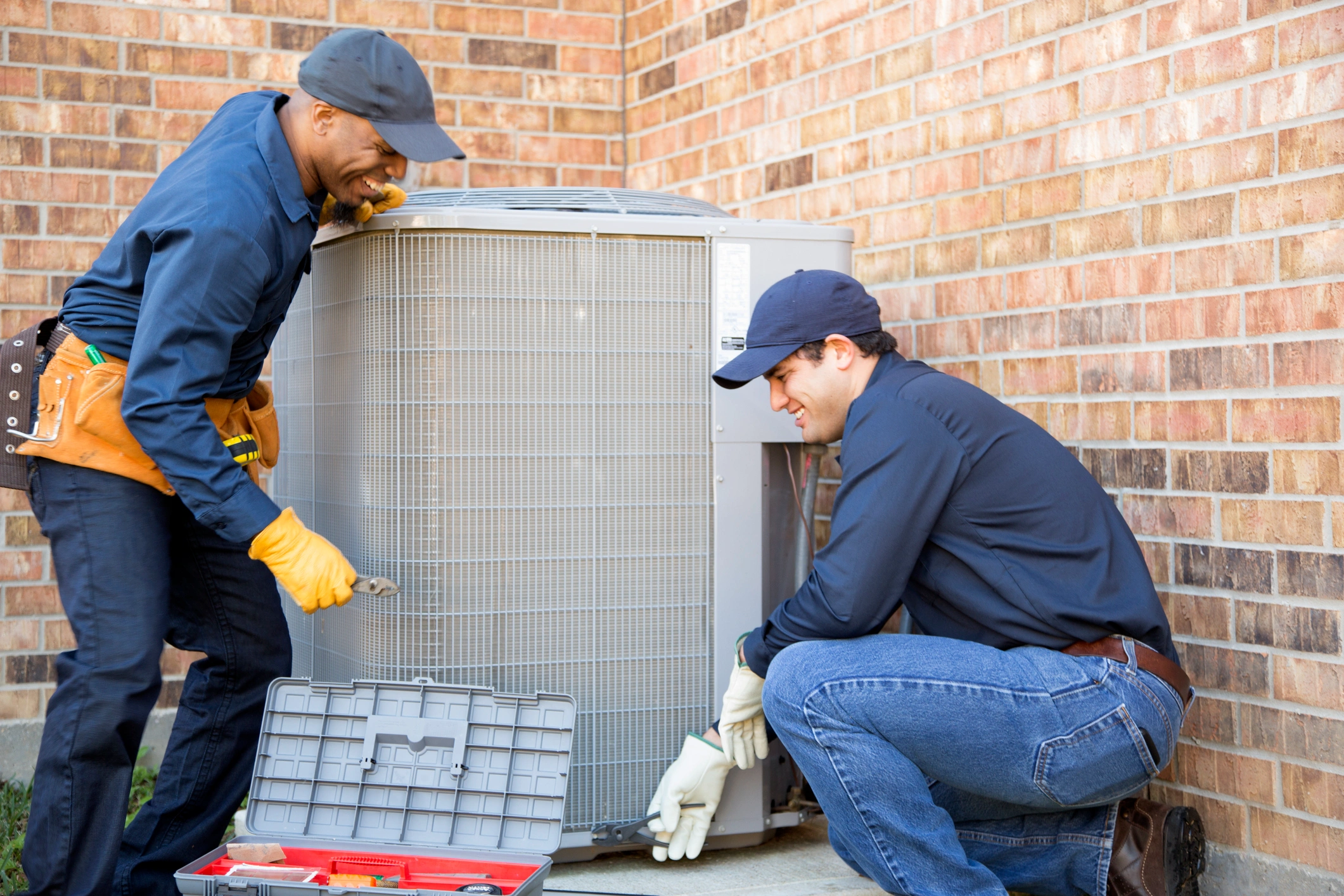 Two technicians working together on an air conditioning unit, tools in hand.